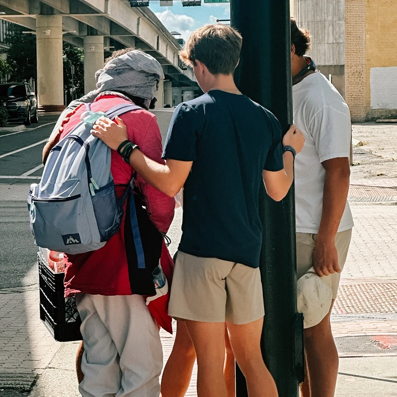 Three people stand on a city sidewalk under an overpass. One young man has his arm around another person wearing a backpack in a gesture of support or prayer.
