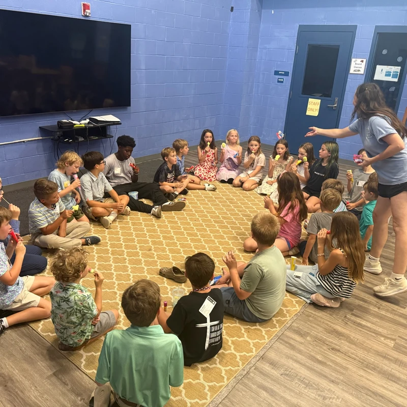 A group of children sit in a circle on a patterned rug, eating ice pops. A female camp counselor stands over them, talking to the group in a blue room.