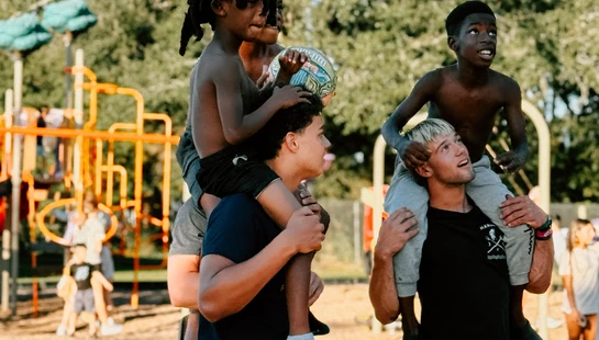 Two young men give two young, shirtless Black boys shoulder rides on a basketball court. A playground and green trees are visible in the sunny background.