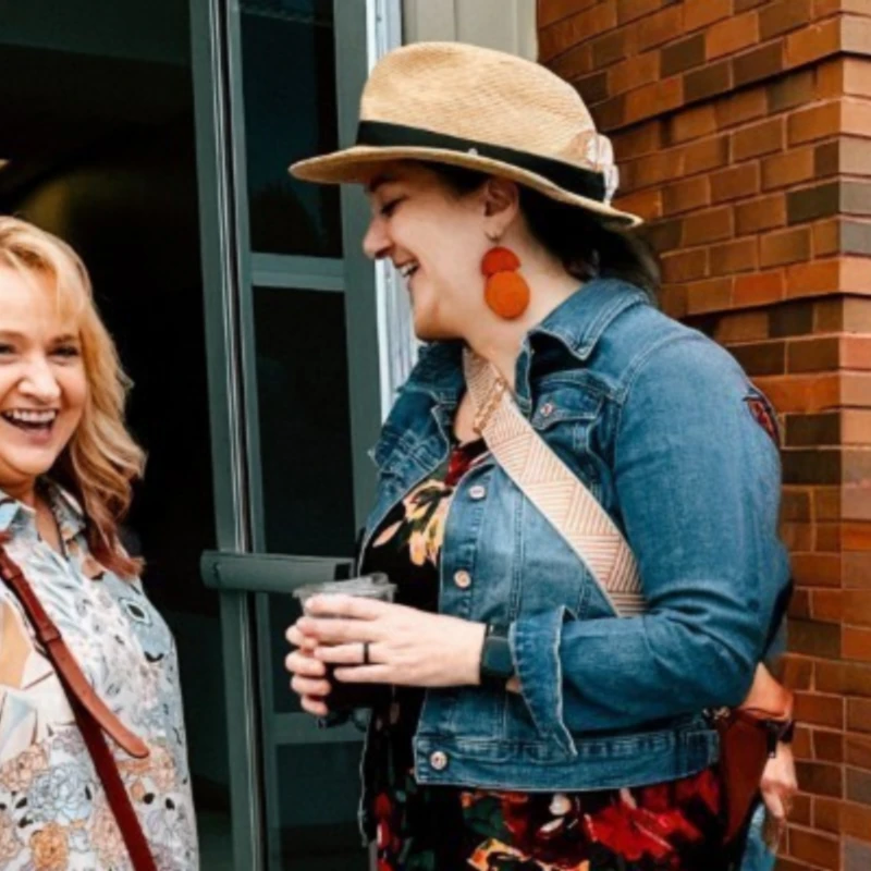 Two women stand outside a brick building, smiling and laughing together. The woman on the right wears a straw hat and holds a drink.