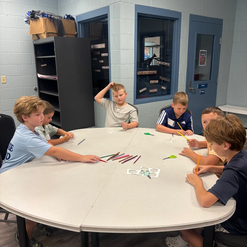 Five young boys sit around a large white table in a classroom, working on an art project with colored pencils and scissors.