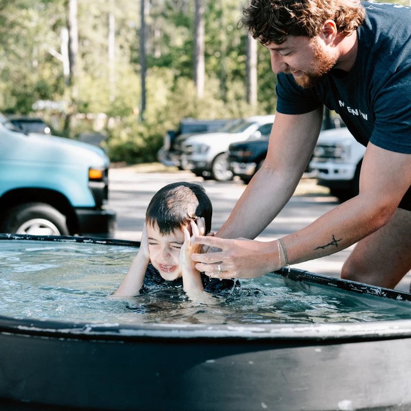 A man with a beard baptizes a young boy in a large black tub of water outdoors. The boy holds his nose and closes his eyes as the man supports him.