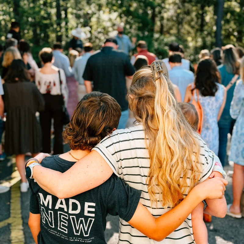From behind, a woman holds a baby and embraces another person wearing a 'Made New' t-shirt. They are part of a large crowd gathered outdoors near a forest.