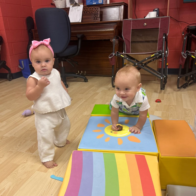A baby girl in a white outfit and pink bow stands, while a baby boy crawls on colorful foam blocks with a sun and rainbow design.