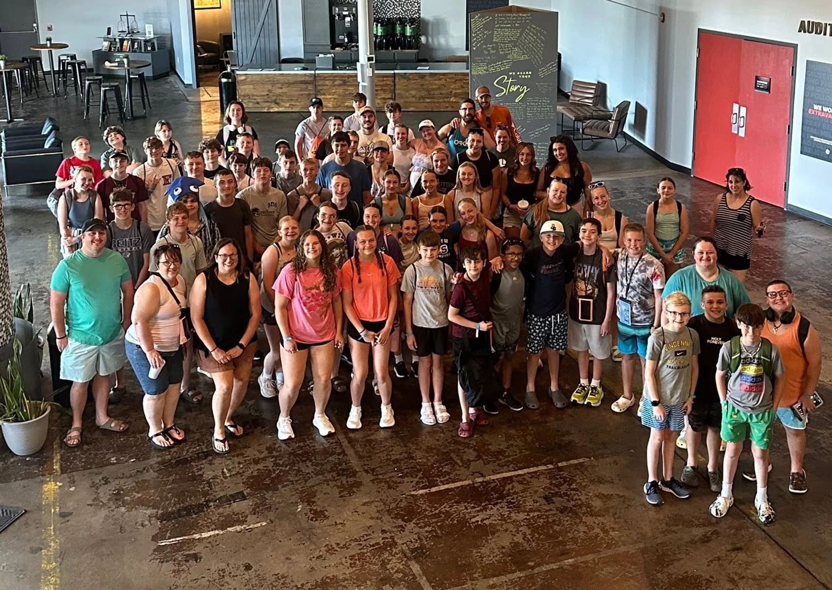 A high-angle group photo of dozens of smiling teenagers and adults standing together on a concrete floor inside a large, modern hall.
