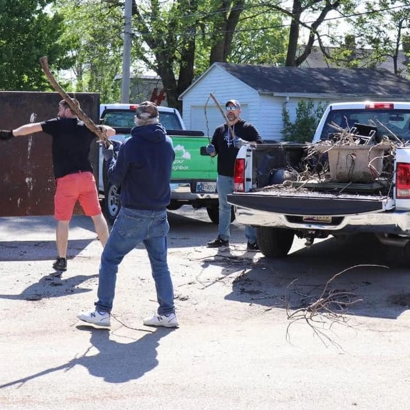 Three men toss large tree branches and other yard waste into a large metal dumpster on a sunny day, with pickup trucks parked nearby.