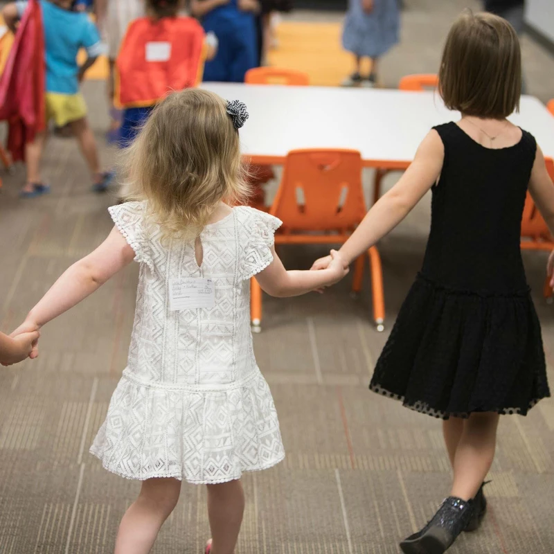 The back of two young girls, one in a white dress and one in a black dress, holding hands as they walk through a room with other children in the background.