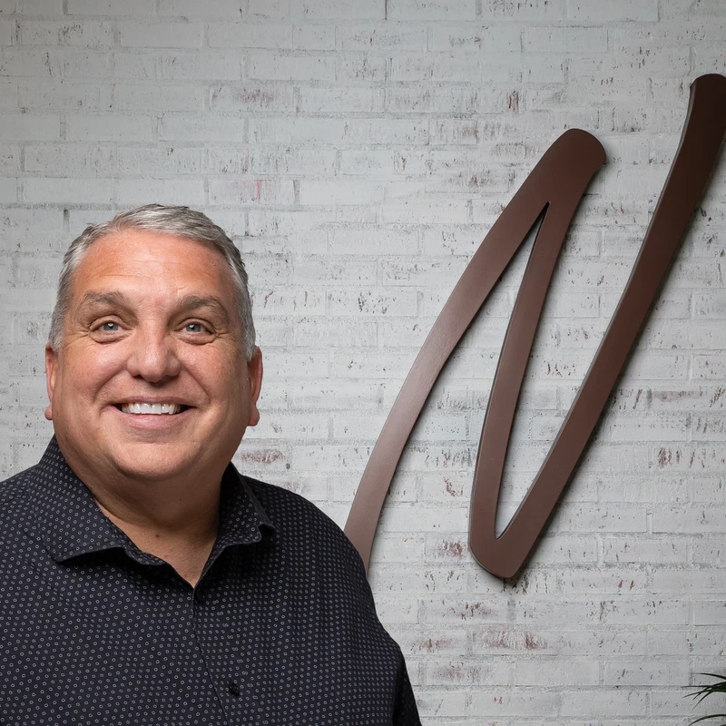 A smiling man with gray hair and a dark shirt stands in front of a white brick wall with a large brown 'M' logo and a plant.