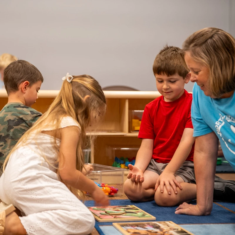 A female teacher and three young children sit on a blue carpet, playing with wooden jigsaw puzzles in a brightly lit classroom.