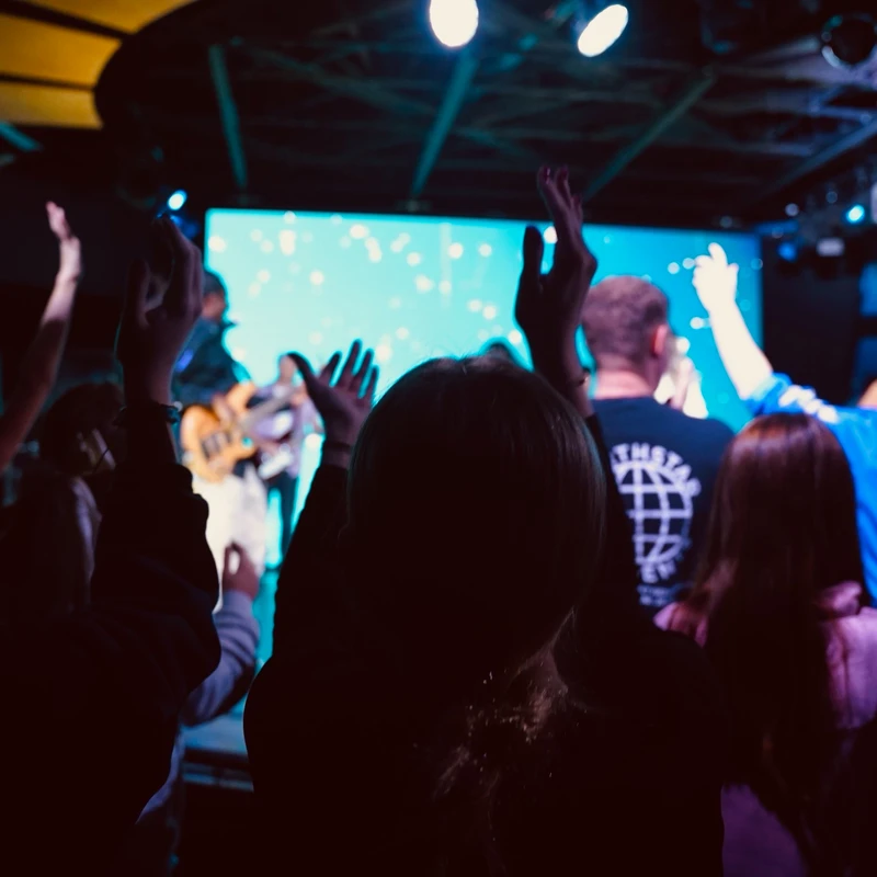 An audience of silhouetted people from behind, raising their hands towards a brightly lit stage with large blue screens during a concert or worship service.