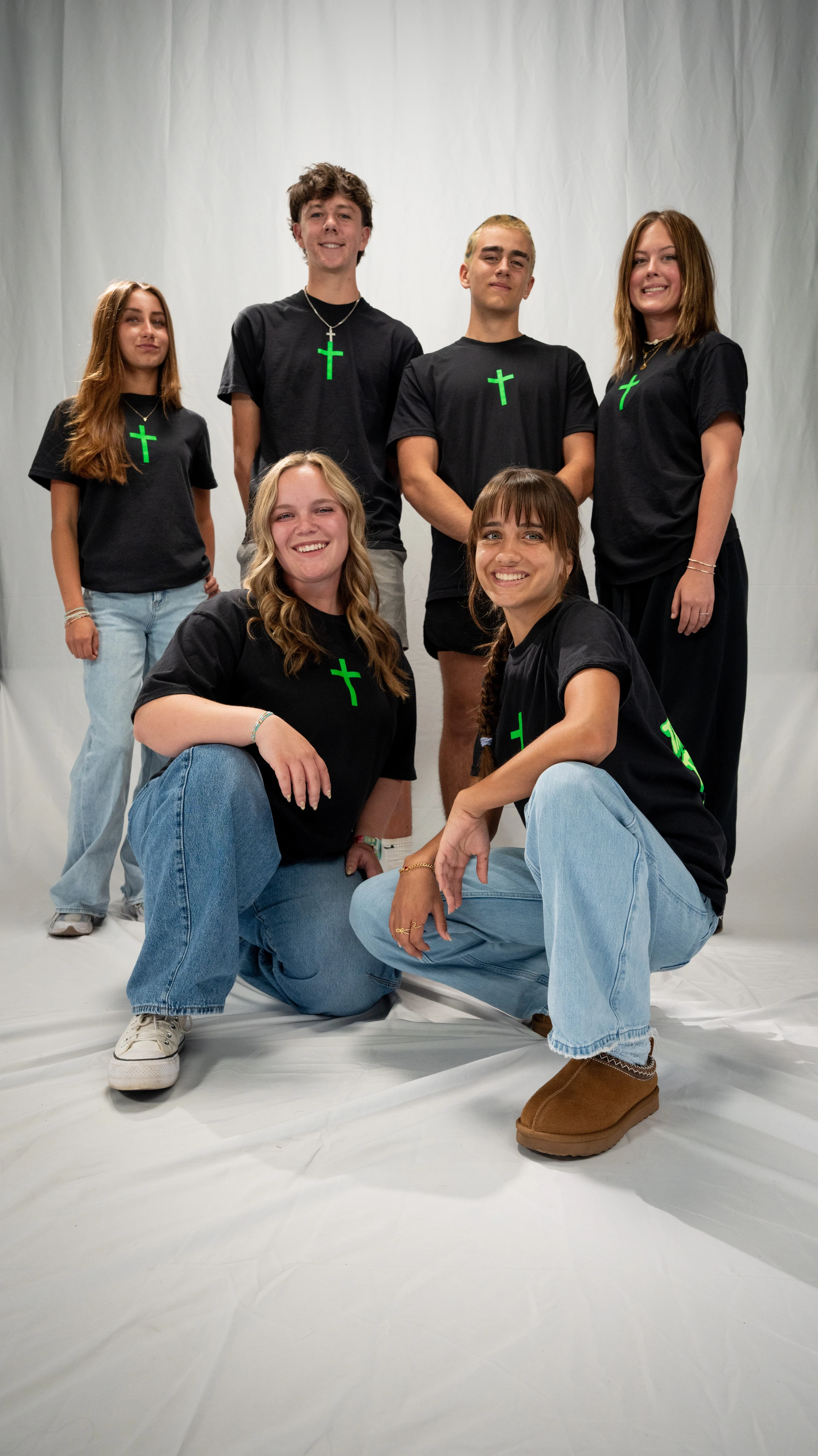 A group of six young people smile for a studio portrait against a white background. They all wear matching black t-shirts with a bright green cross.