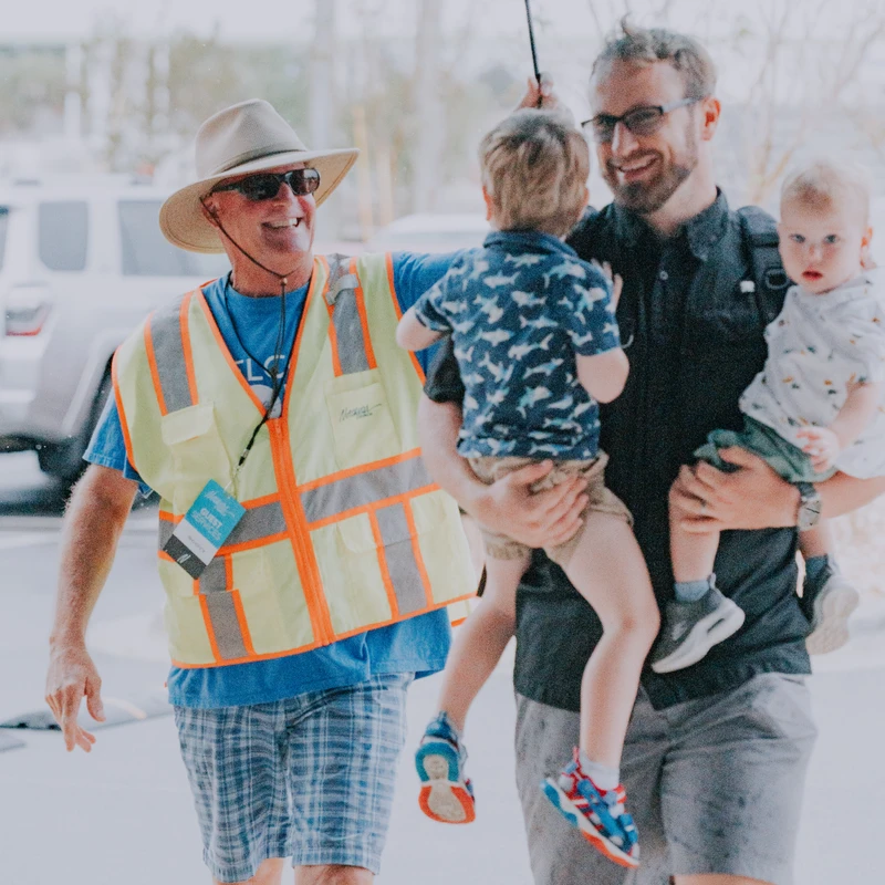 A smiling volunteer in a safety vest holds an umbrella over a father carrying his two young children as they walk together in the rain.