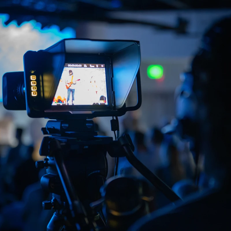 A close-up of a video camera monitor showing a guitarist on stage during a concert. The scene is lit with cool blue light, focusing on the equipment.