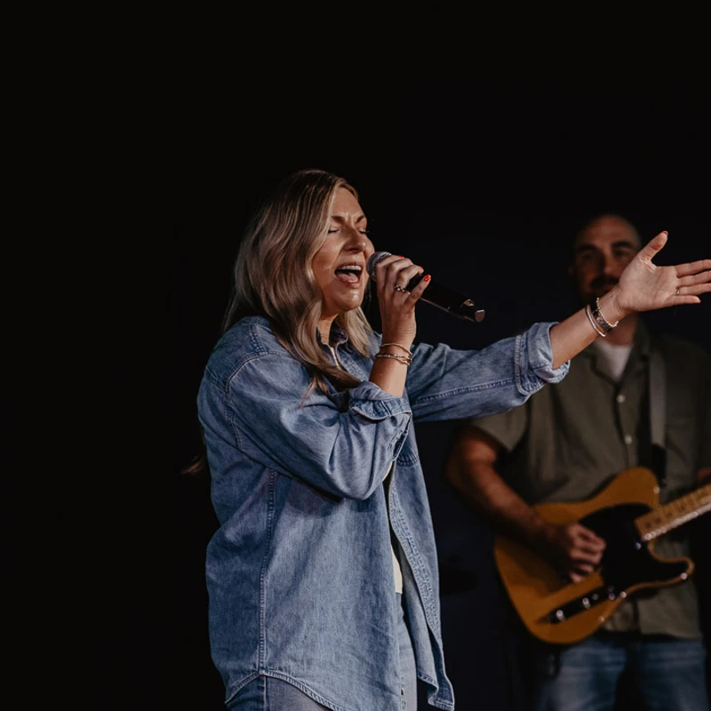 A blonde woman in a denim shirt sings into a microphone with her eyes closed and one arm outstretched. A man plays guitar in the dark background.