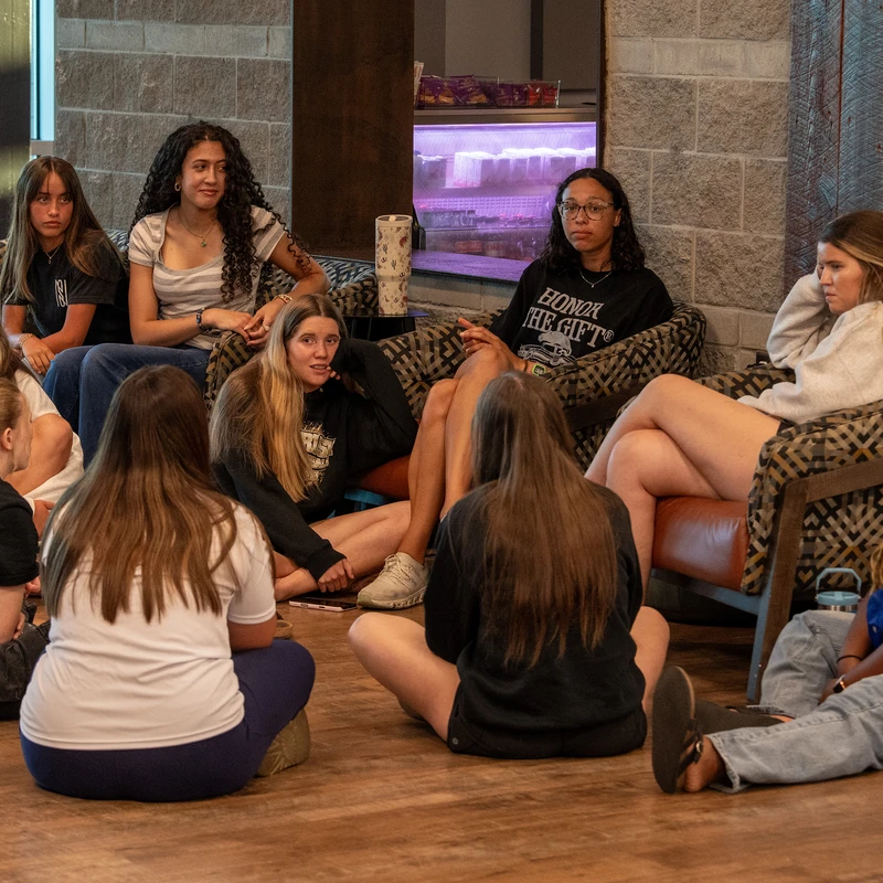 A diverse group of young women sit in a circle on chairs and the floor, attentively listening and engaging in a conversation in a common area.