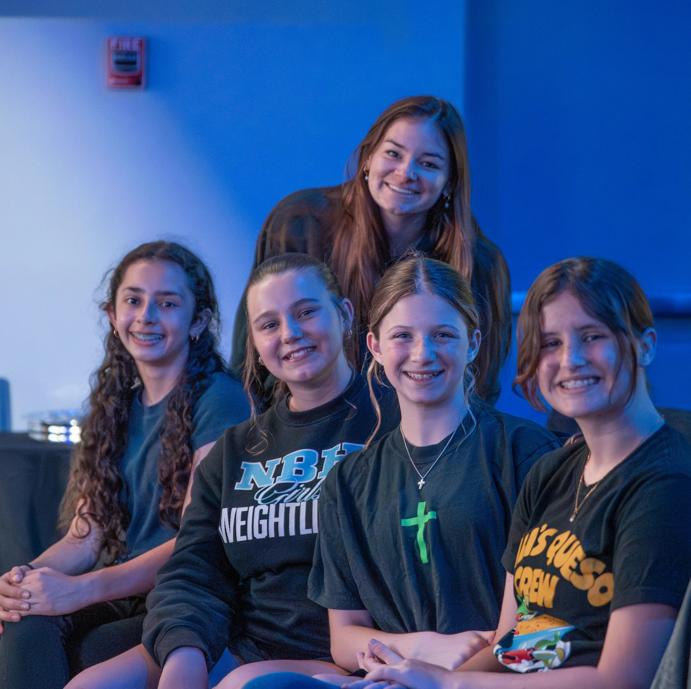 A group of five young women sit and stand together, smiling for a group portrait in front of a solid wall with blue lighting.