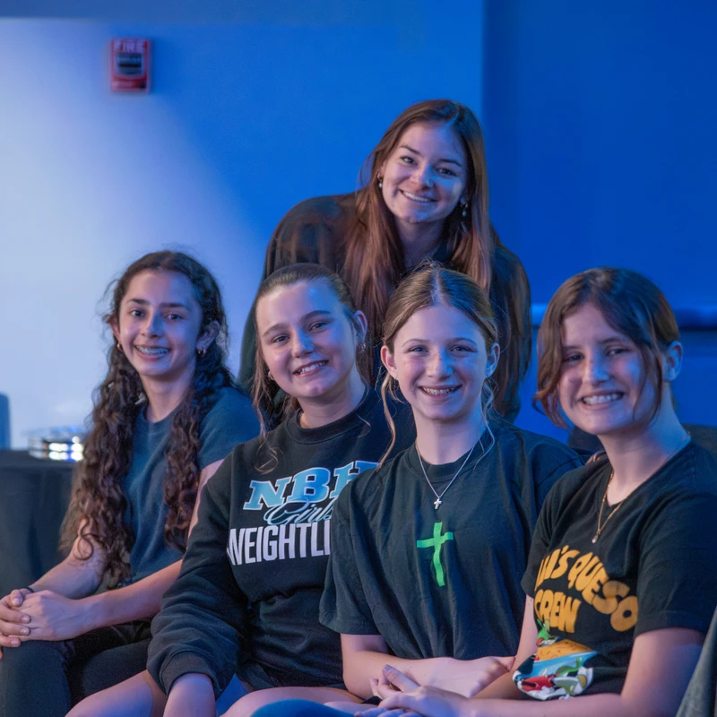 A group of five young women smile for a portrait against a blue-lit wall. Four are sitting in a row and one stands behind the middle two.
