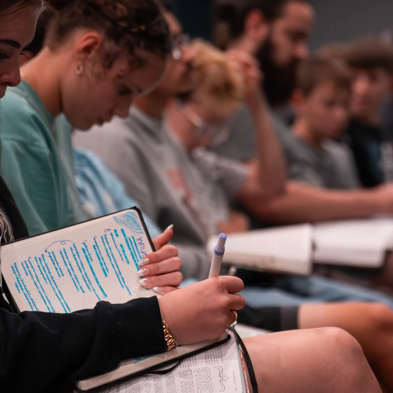 A close-up of a student taking notes in blue ink in a journal during a lecture, with other attendees sitting in rows in the background.
