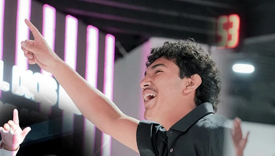 A young man with curly dark hair smiles with his mouth open, pointing one finger up towards the ceiling at an indoor event with pink lights.