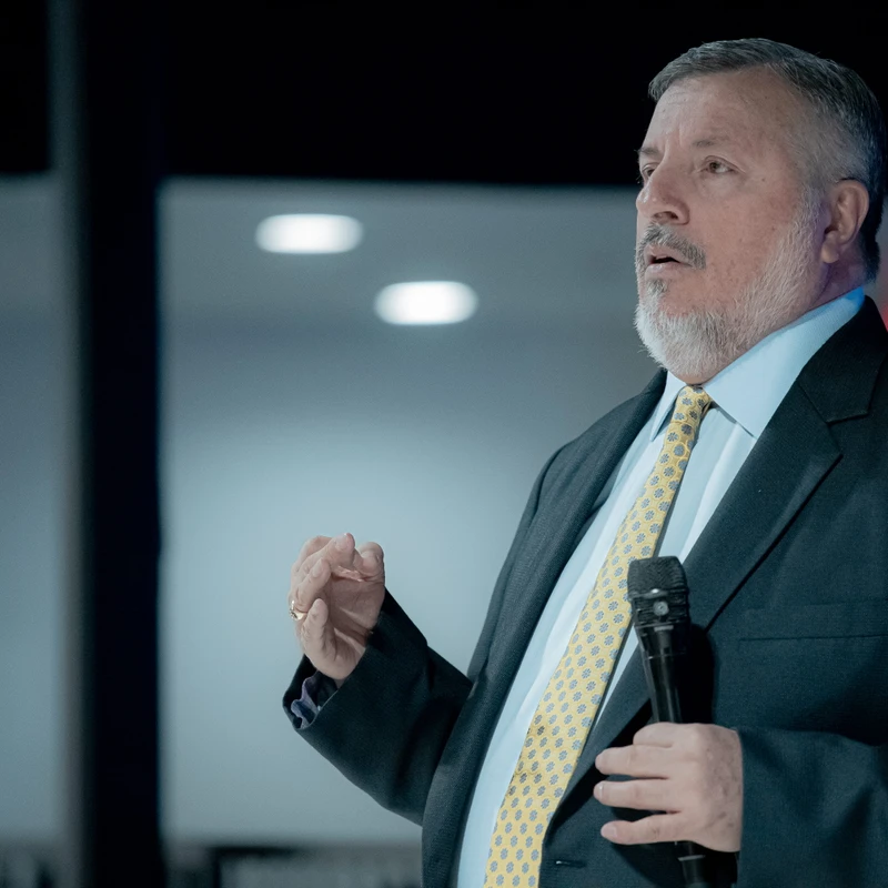 A middle-aged man with gray hair and a beard, wearing a dark suit and patterned yellow tie, holds a microphone and gestures while speaking.