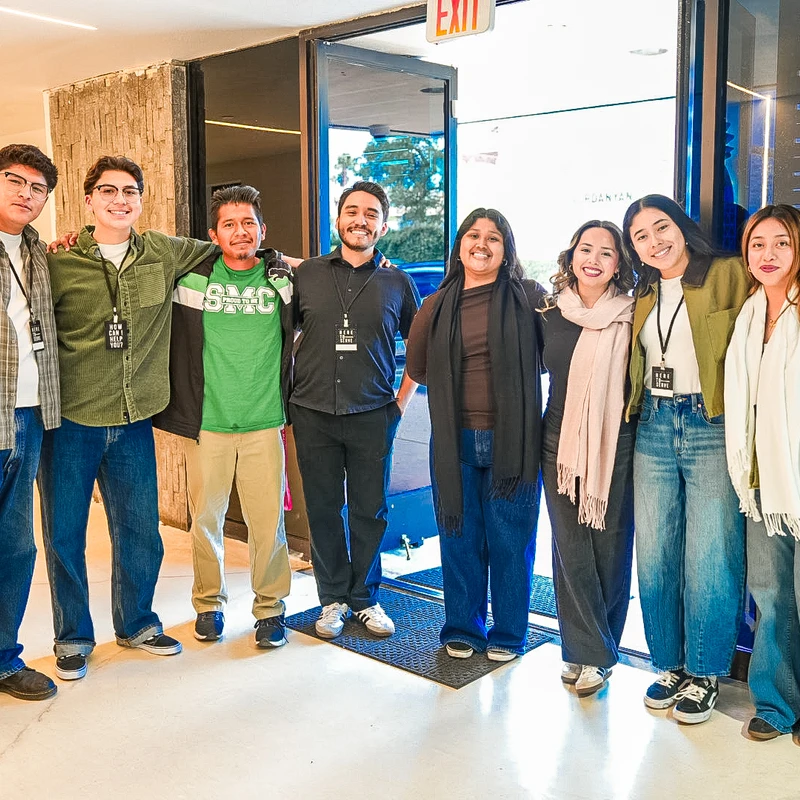 A diverse group of eight smiling people stands together with arms around each other in the brightly lit doorway of a building.
