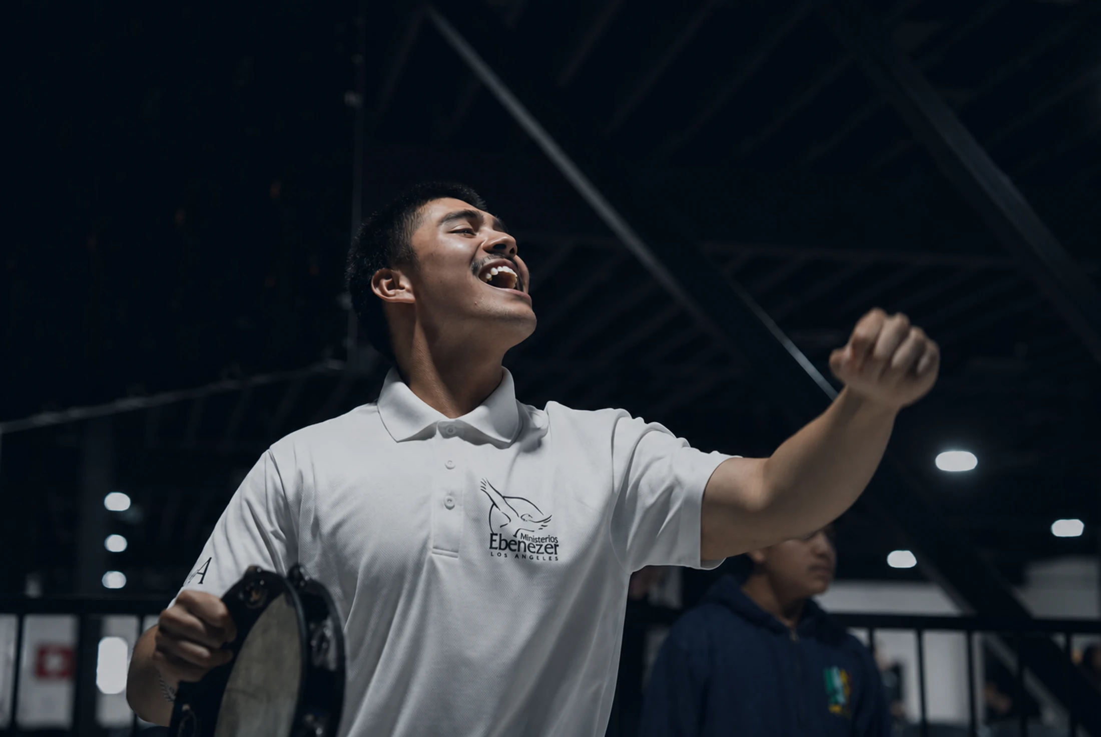 A young man in a white polo shirt sings with passion, looking upwards with his mouth open. He holds a tambourine in one hand and raises a fist in the air.