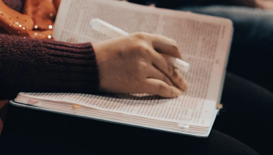 A close-up of a person's hand holding a white pen over an open book on their lap, with other people seated in the blurry background.