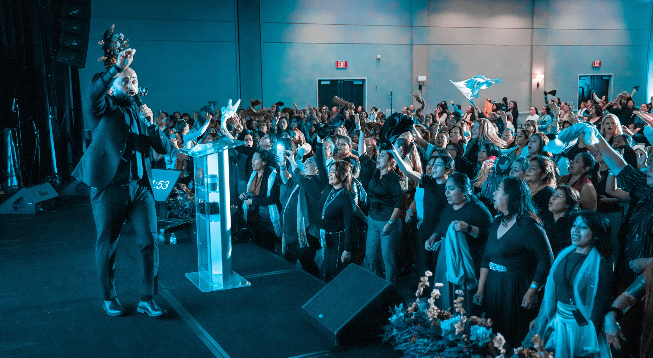 A male speaker in a suit on a blue-lit stage gestures to an enthusiastic crowd of women who are cheering and raising their hands in the air.