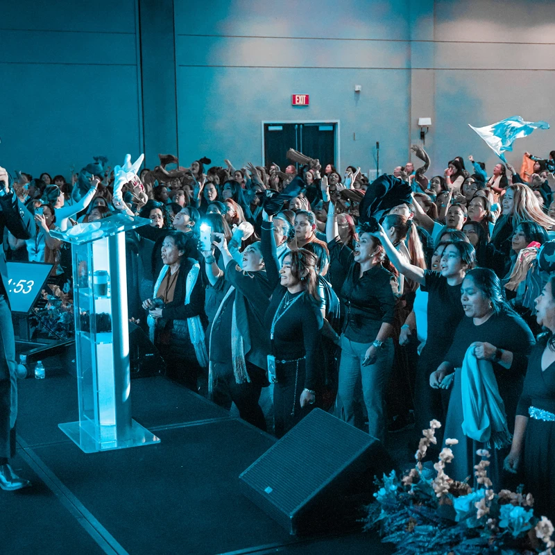 A male speaker in a suit on a blue-lit stage gestures to an enthusiastic crowd of women who are cheering and raising their hands in the air.