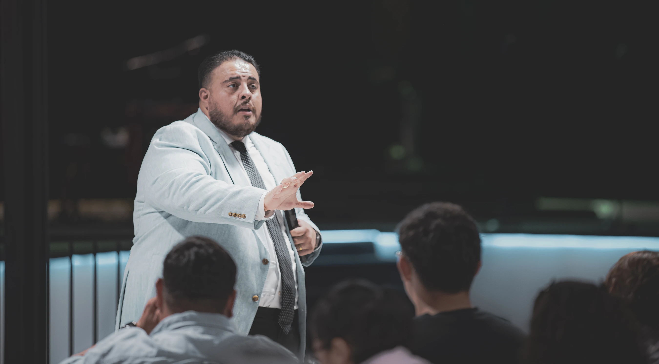 A man with a beard, wearing a light-colored suit and tie, stands and speaks to a seated audience, gesturing with his hand. The background is dark.
