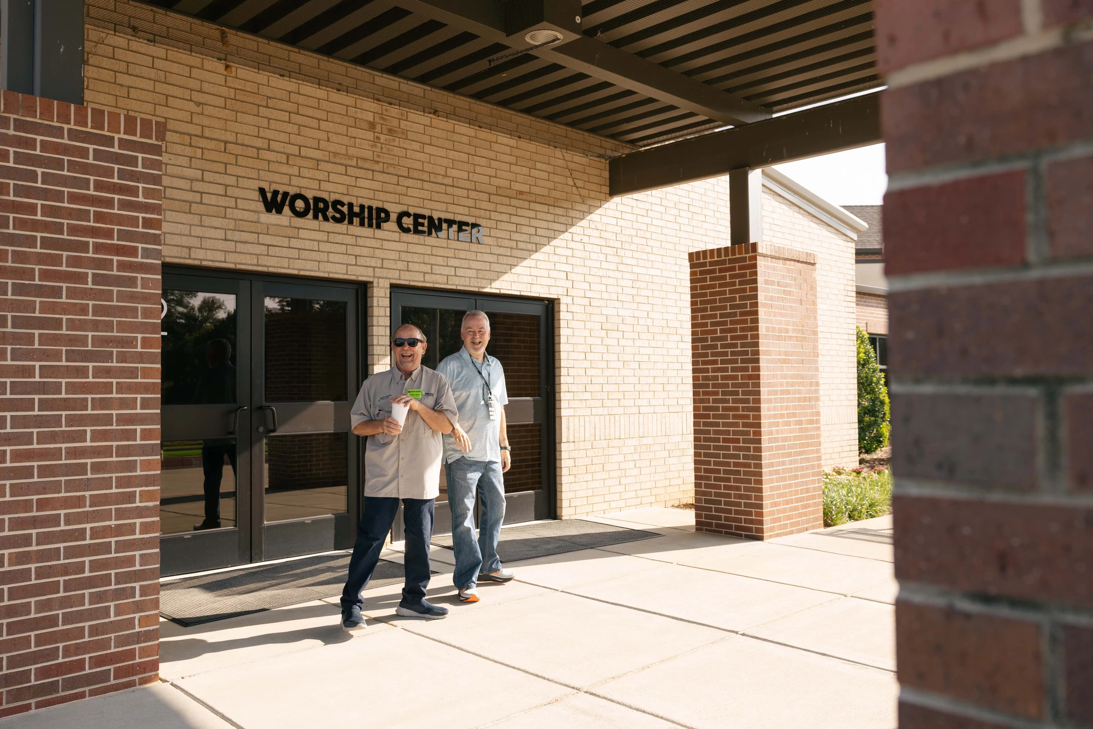 Two smiling men walk out from the entrance of a brick building labeled 'Worship Center' on a sunny day. One man wears sunglasses and holds a cup.