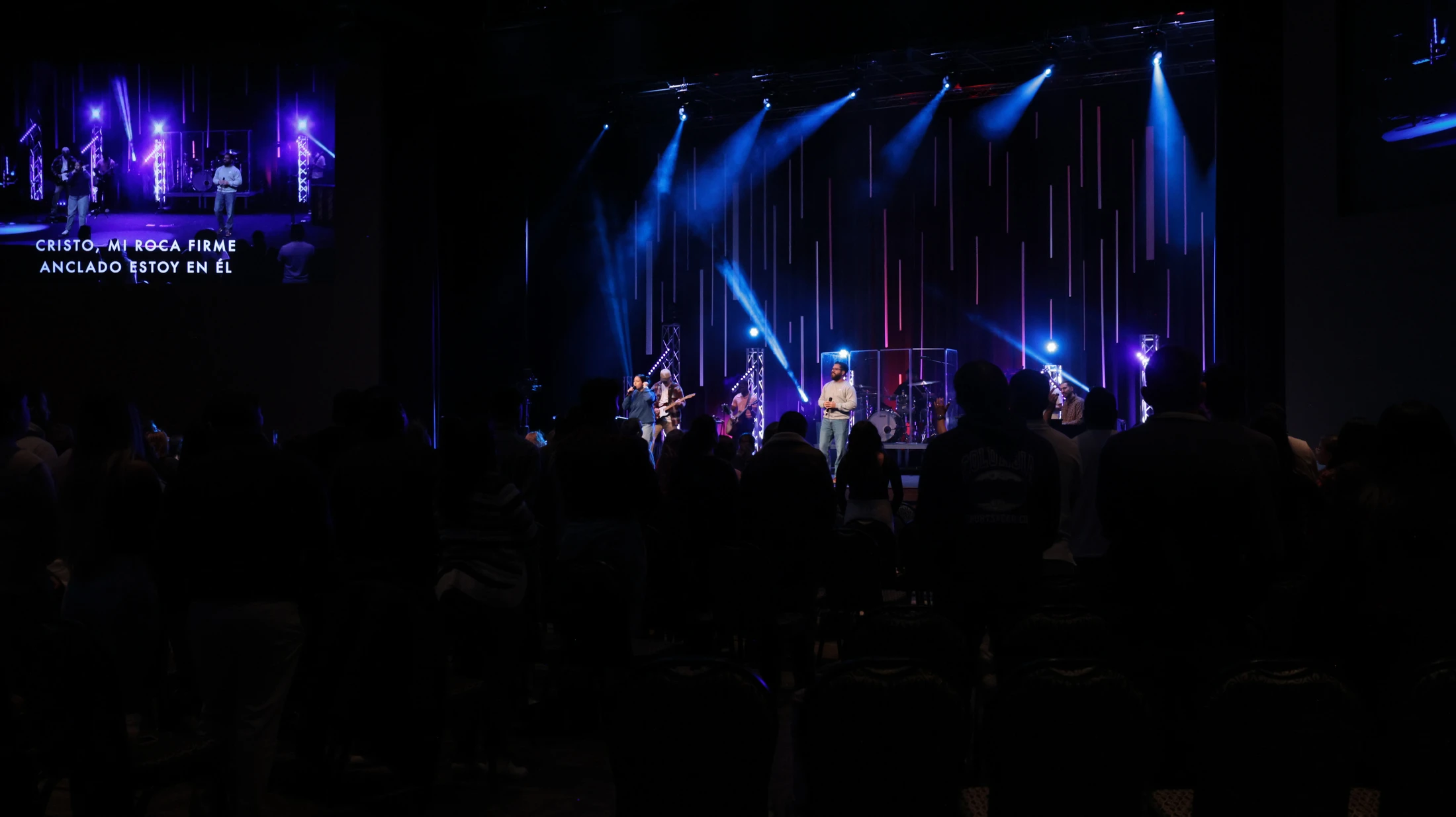 A silhouetted audience watches a worship band perform on a stage with dramatic blue lighting. A screen to the left displays lyrics in Spanish.