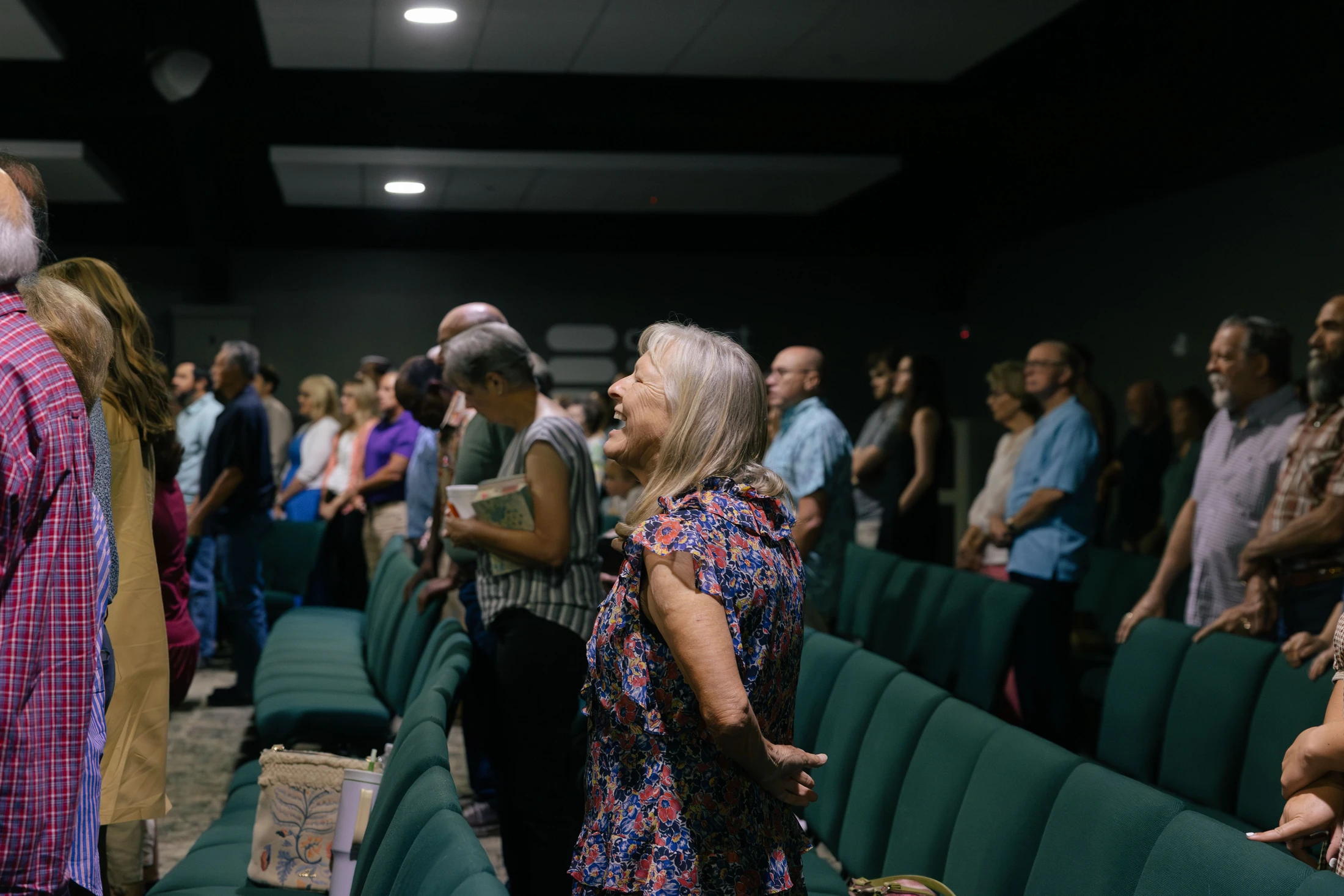 A woman with blonde hair in a floral dress looks up and smiles broadly while standing among a congregation in an auditorium with green seats.