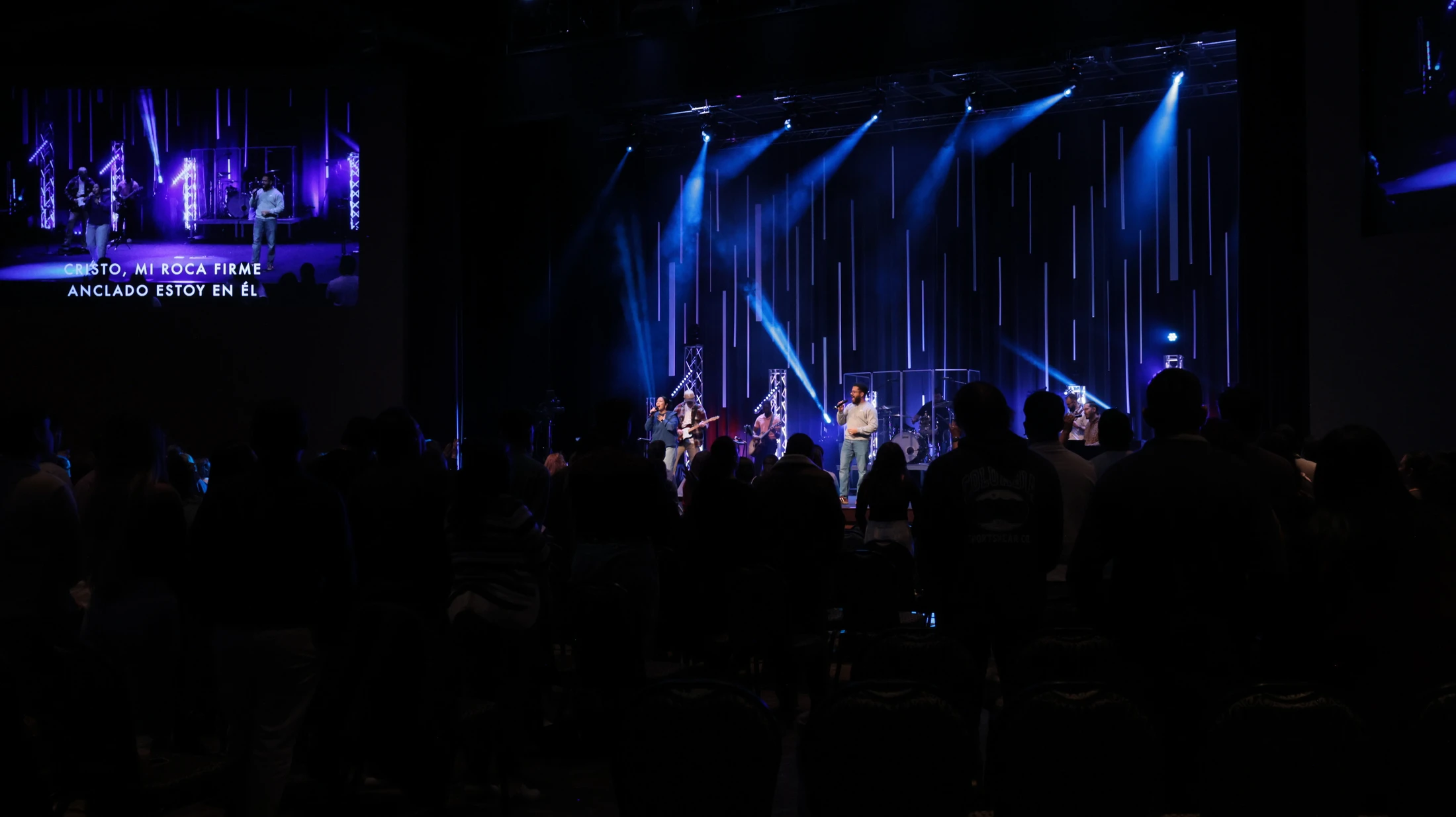 A silhouetted audience watches a worship band perform on a stage illuminated by blue spotlights. A screen to the side shows lyrics in Spanish.