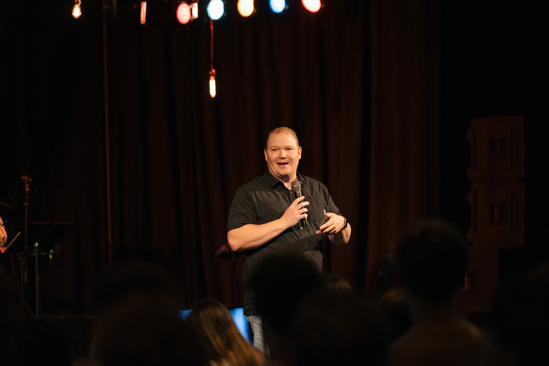 A man in a black shirt stands on a dimly lit stage, holding a microphone and speaking to an audience. Stage lights hang above him in front of a dark curtain.