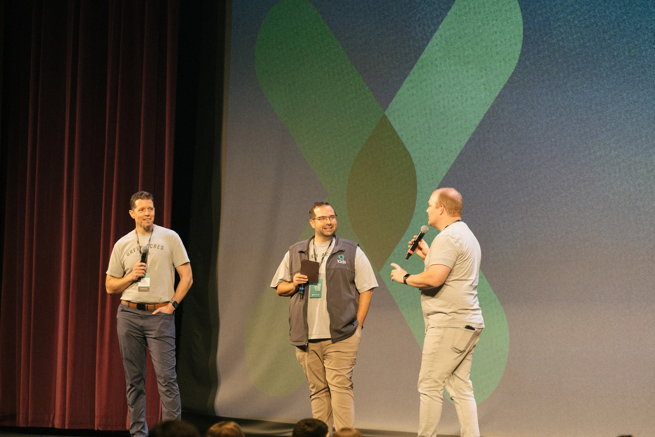 Three men stand on a stage, speaking into microphones. They are in front of a screen with a large green 'X' graphic and a red curtain to the side.