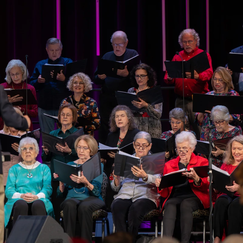 A choir of older adults, men and women, sing from music binders while seated on a dimly lit stage. A conductor in the foreground leads them.
