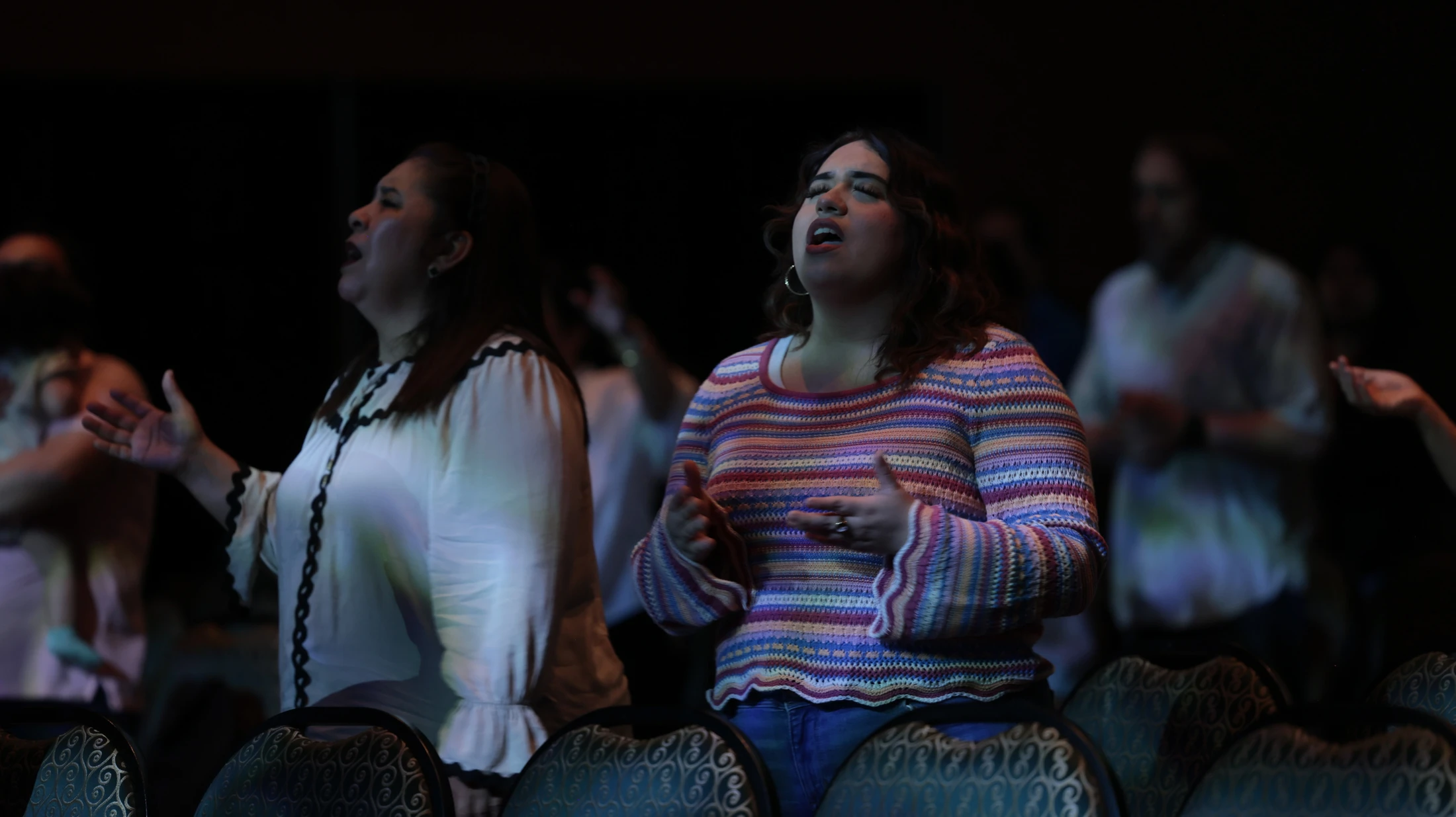 Two women stand in a dimly lit room with a congregation, singing with their eyes closed and hands raised in an act of worship or praise.