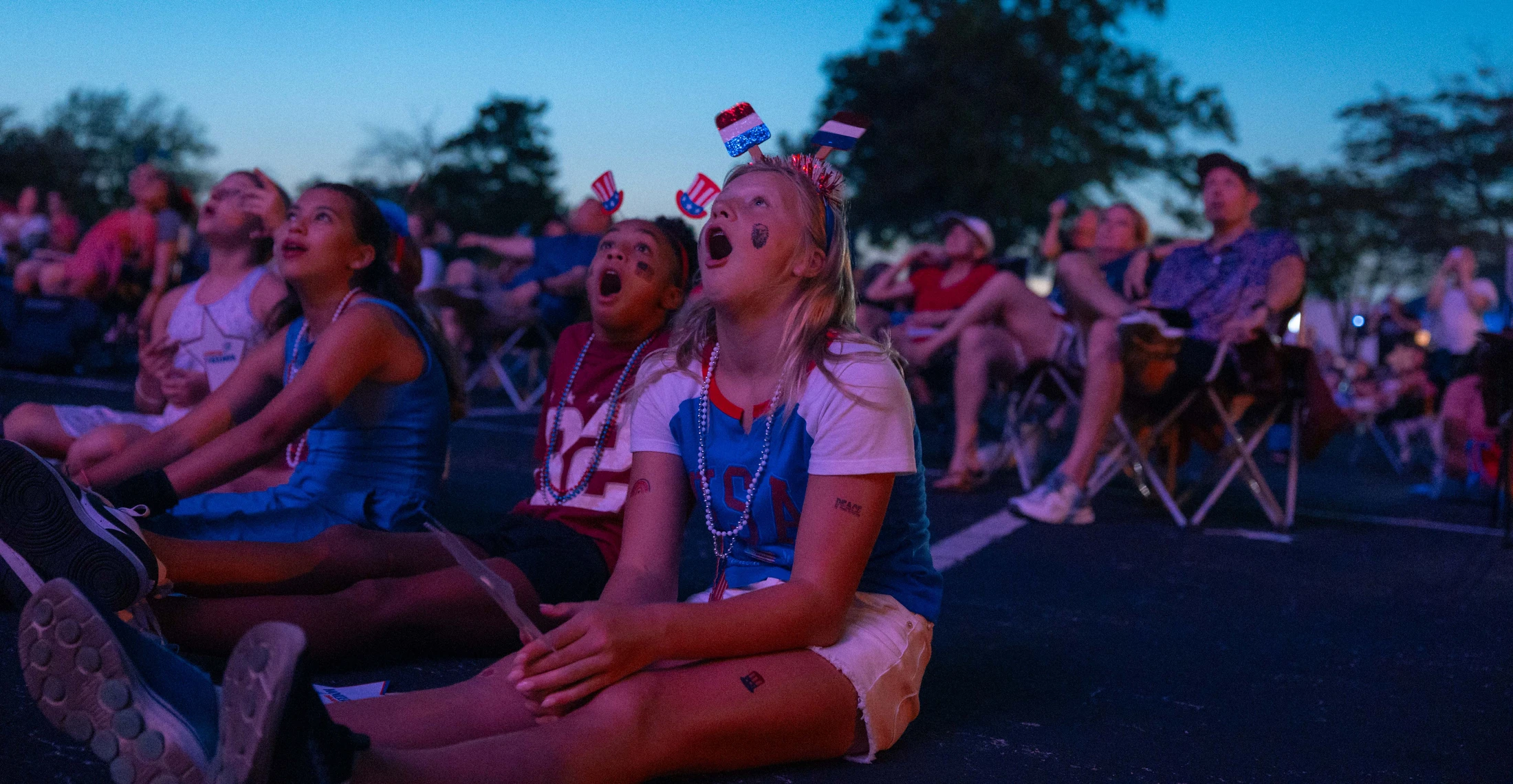 A group of children in patriotic attire sit on the ground at dusk, looking up with expressions of wonder and excitement at a fireworks show.