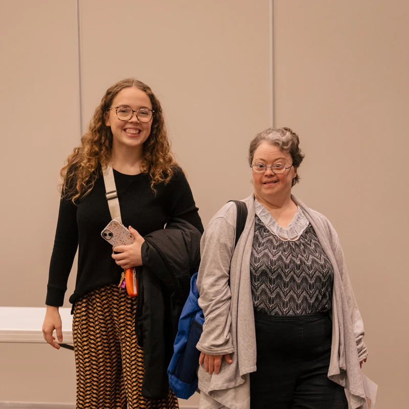 A young woman with curly hair and glasses stands smiling next to an older woman with Down syndrome, also smiling. They are posing together indoors.