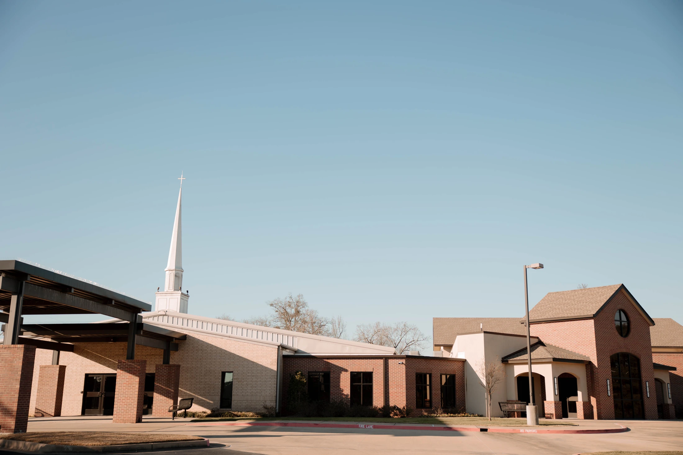 A modern brick church with a covered entrance and a tall white steeple in the background, seen from the parking lot on a sunny day.