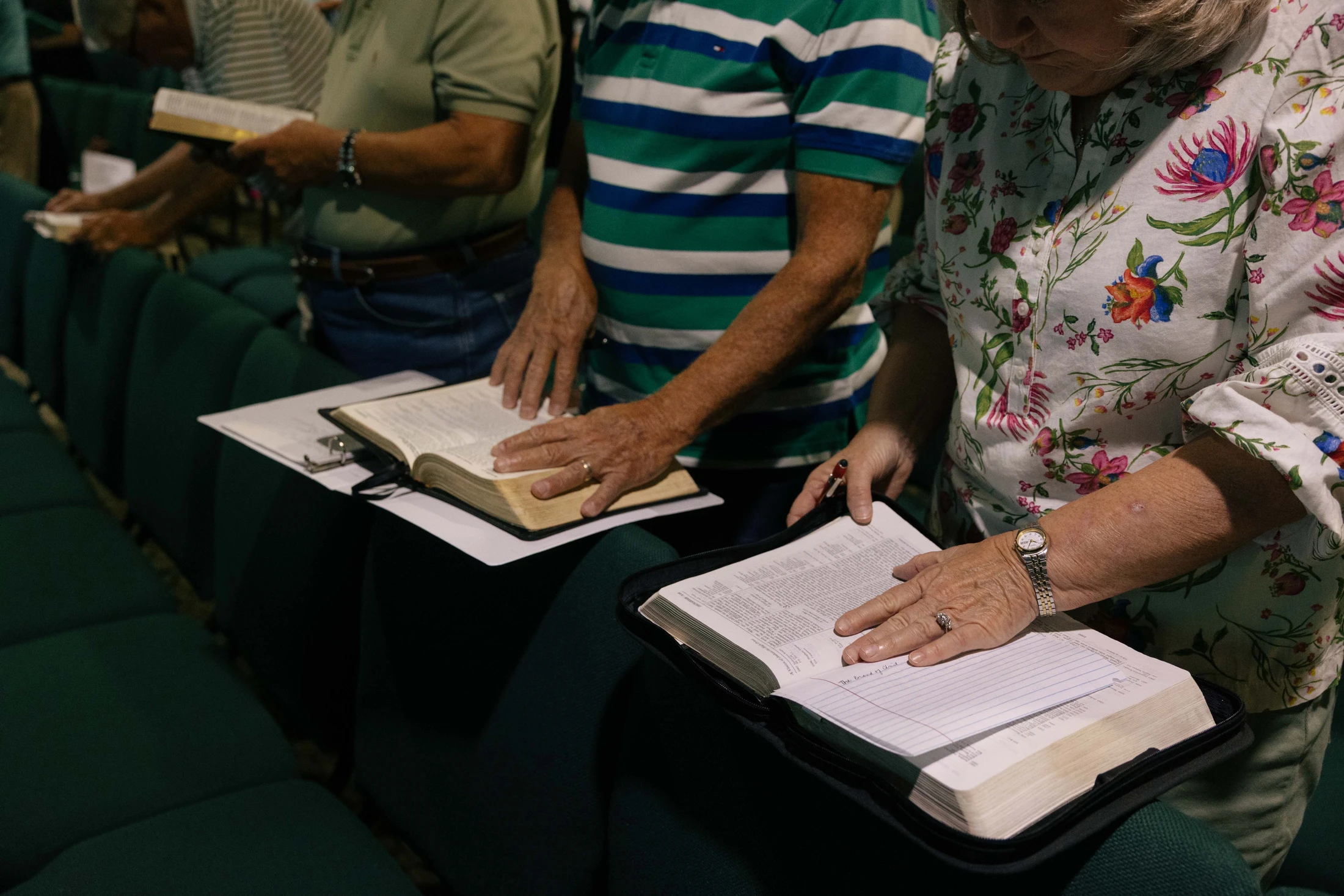 Several people stand in a row in a church, each holding an open Bible. The focus is on their hands as they follow along with the text during worship or study.