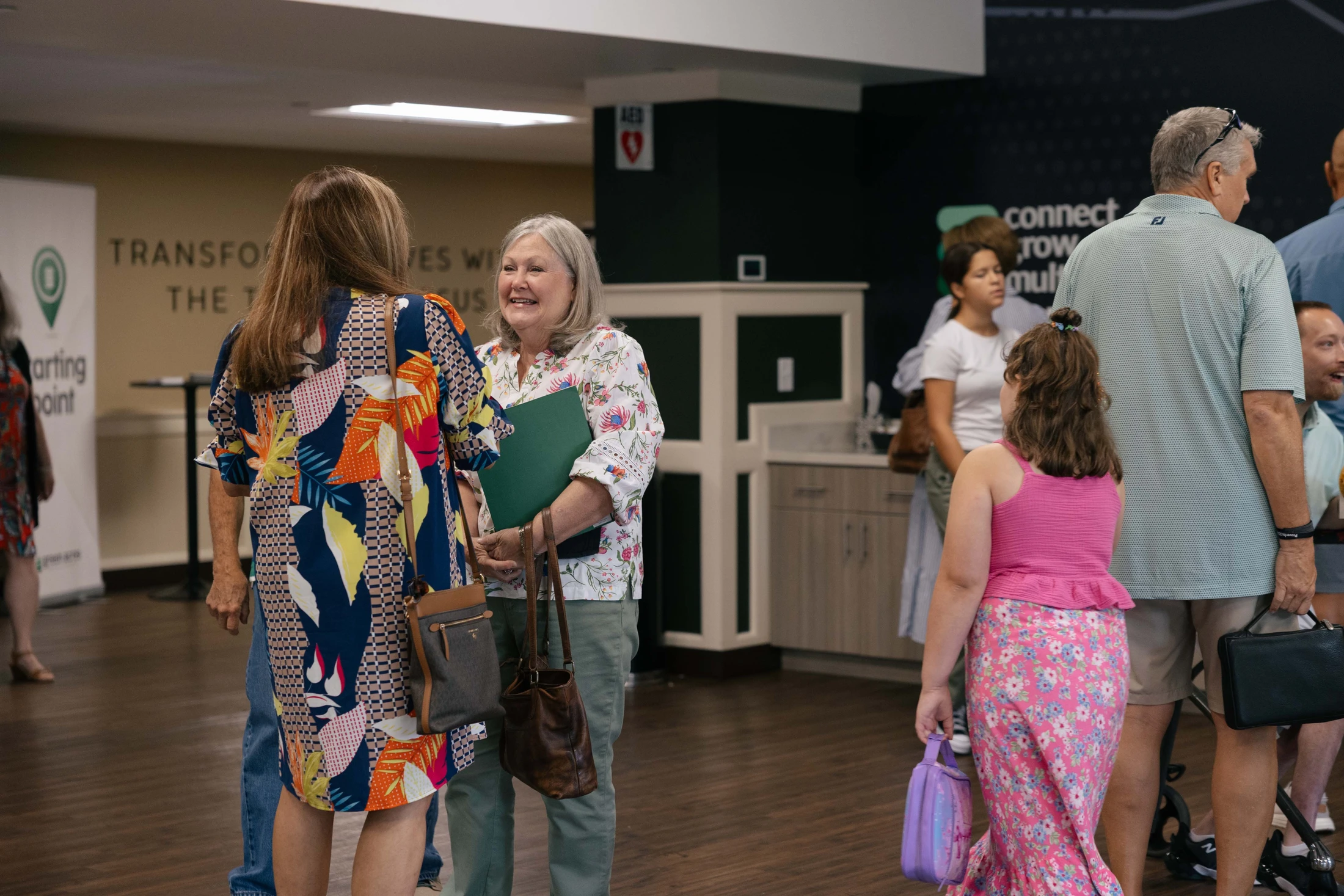 Two women smile and talk in a busy room. One wears a colorful dress, the other a floral blouse while holding a folder. Others mingle in the background.