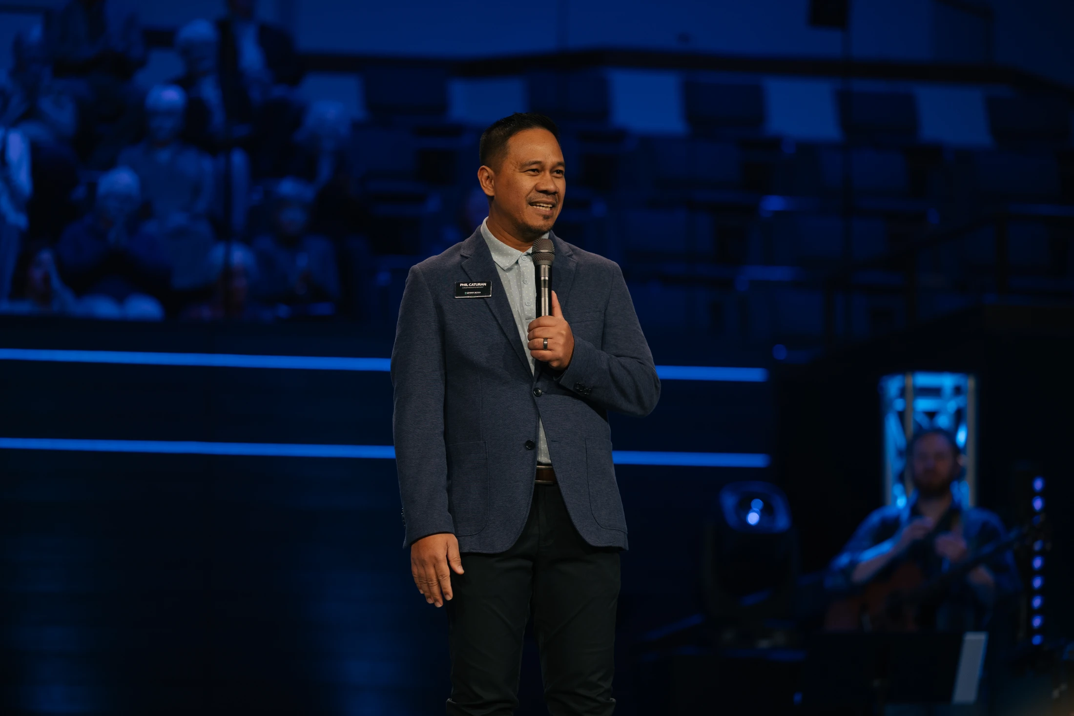 A smiling man in a blue blazer and black pants stands on a dark stage with blue lighting, holding a microphone and speaking to an audience.