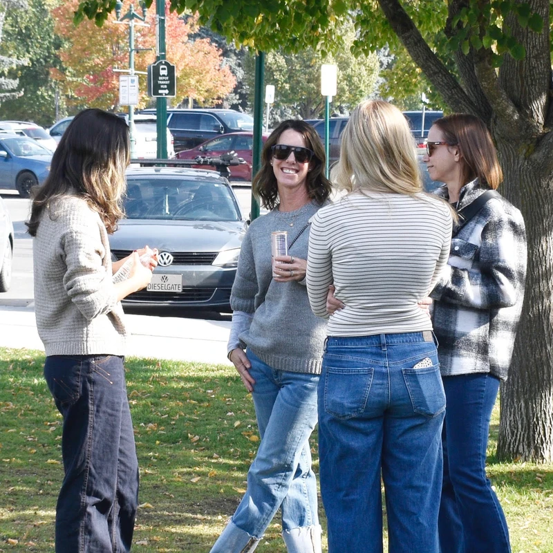 Four women in casual sweaters and jeans stand on a grassy area under a tree, talking and smiling on a sunny day. Parked cars are visible in the background.