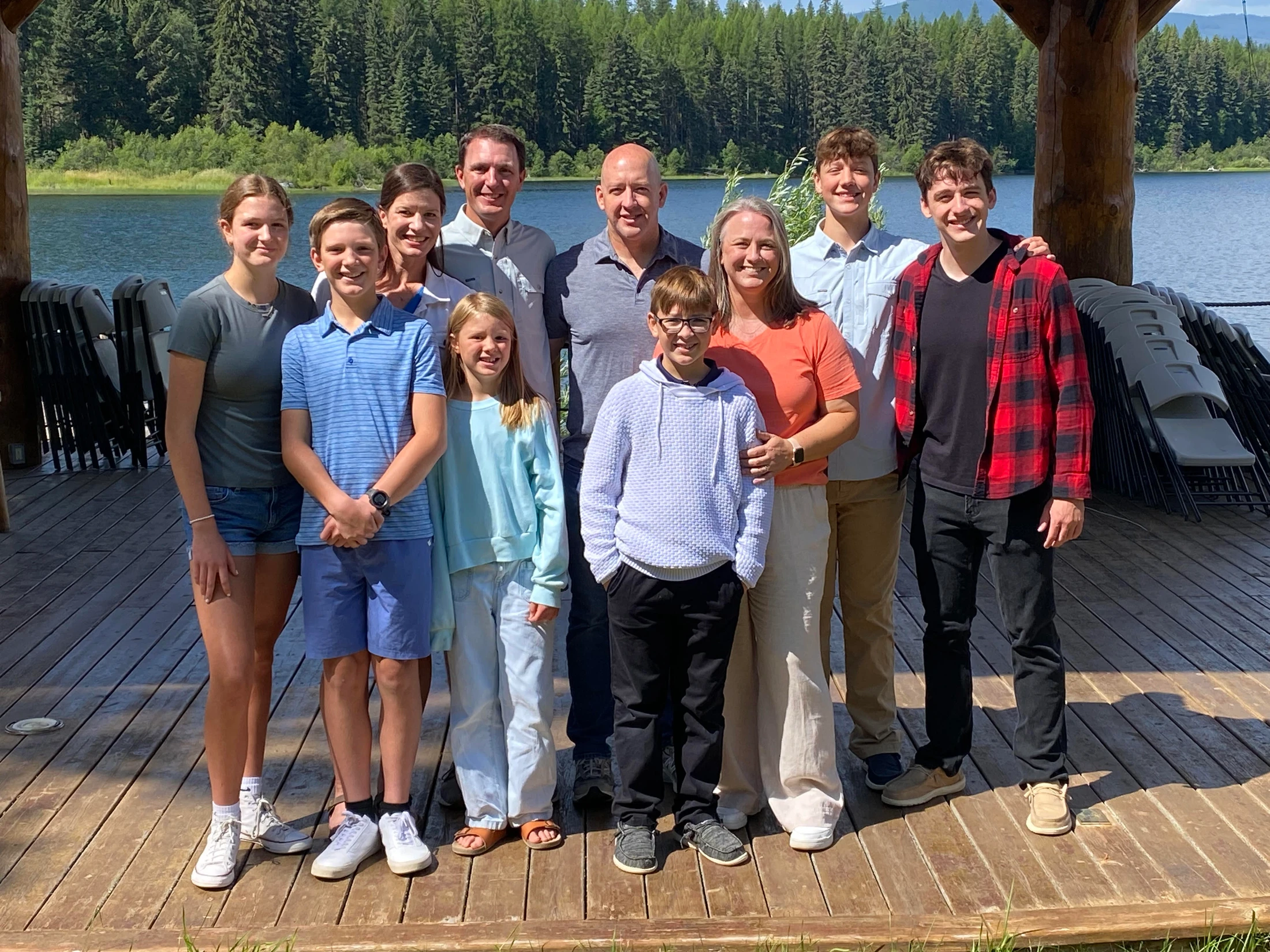 A large family of ten people smiles for a photo on a wooden dock. In the background is a calm lake and a dense forest under a clear sky.
