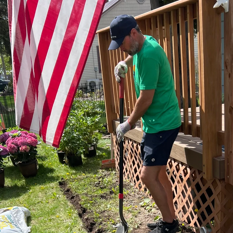 A man in a green shirt and cap digs a trench with a shovel in a grassy yard next to a deck. An American flag and potted plants are nearby.