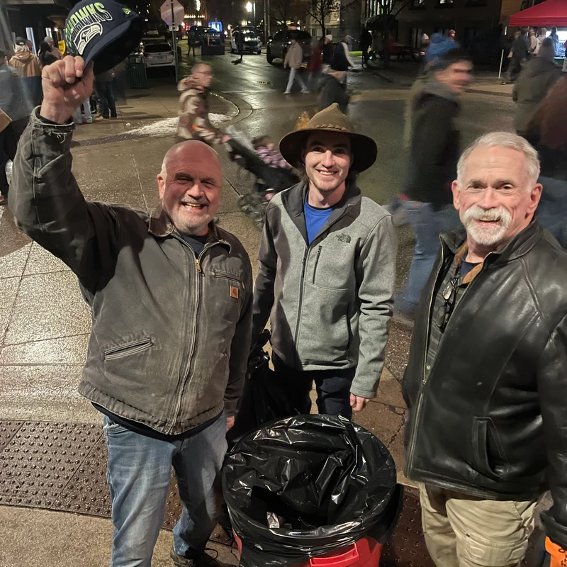 Three smiling men pose for a photo on a city sidewalk at night. One man holds up a Seattle Seahawks hat. A red trash can is in front of them.