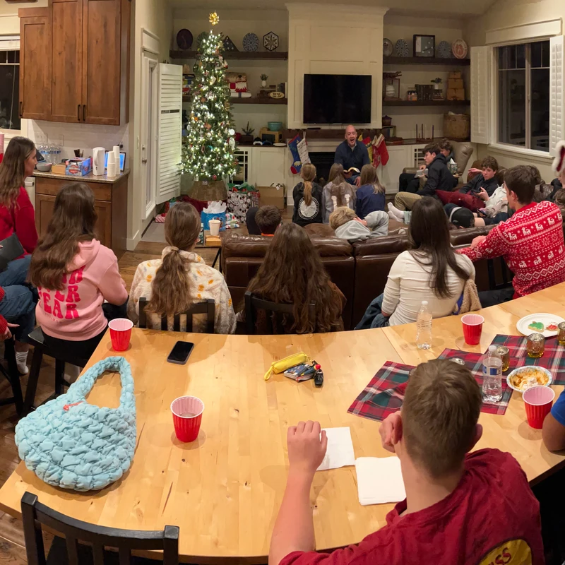 A panoramic photo of a large group of people in a home at Christmas, gathered around a man reading a story aloud by a decorated, lit Christmas tree.
