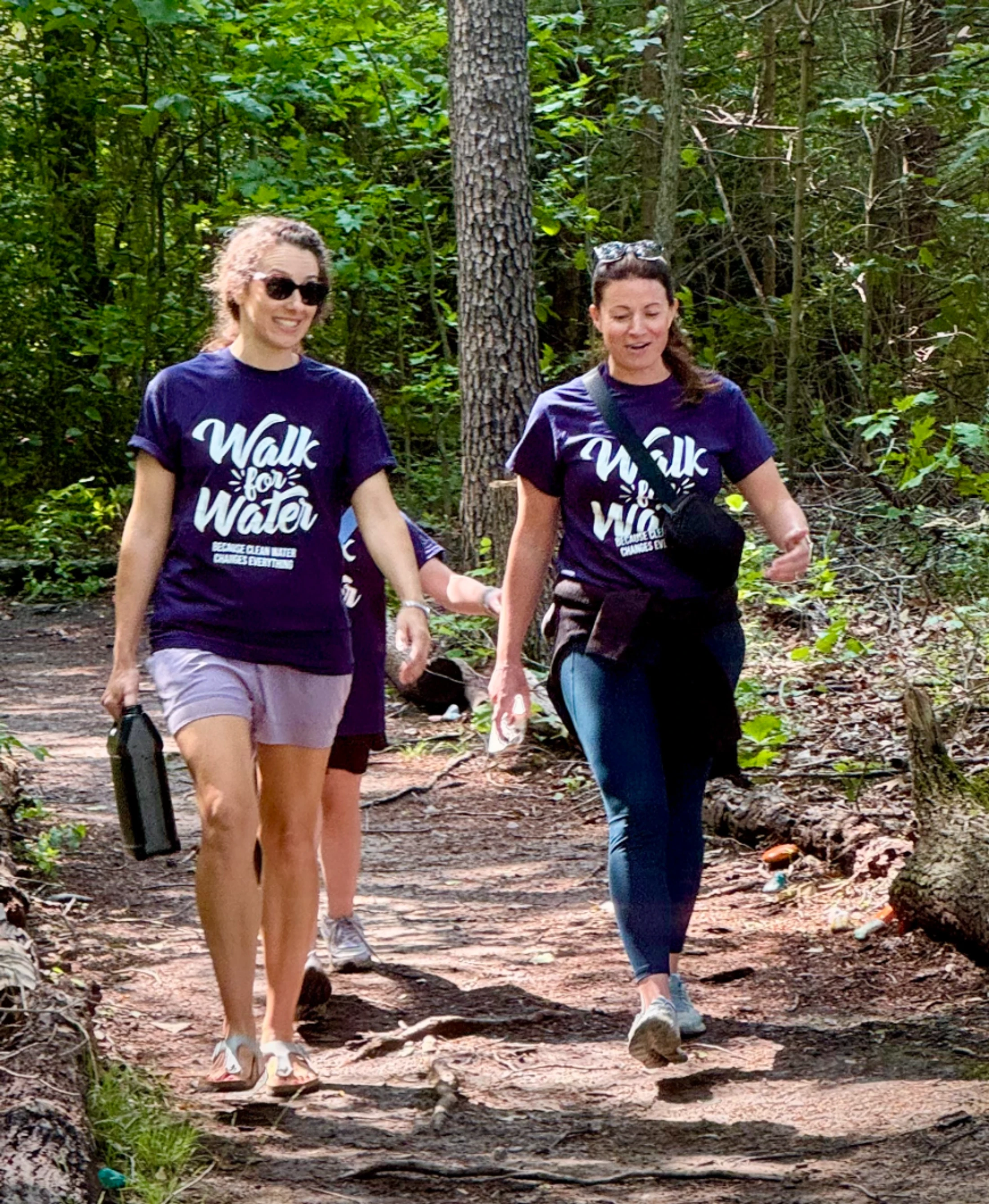 Two women wearing purple 'Walk for Water' t-shirts walk along a dirt path in a sunny forest, participating in a charity event.